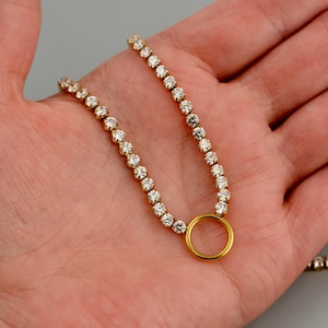 May include: A gold-toned chain necklace featuring a continuous row of small, clear, round-cut stones set in metal prongs, terminating in a smooth, circular gold-toned ring pendant that connects the two ends of the sparkling stone-studded chain.