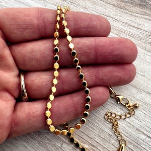May include: A gold-colored necklace with a delicate design of small, flat, round beads. Some beads are black. The necklace is held in a hand, and the clasp and extender chain are visible. The background is a light-colored wooden surface.