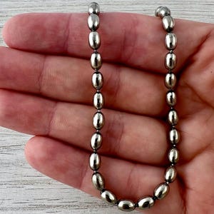 May include: A silver-toned necklace composed of oval-shaped beads. The beads are strung together to form a continuous loop. The necklace is held in a hand, showcasing its design and metallic sheen. The beads are approximately 0.5 inches in length.