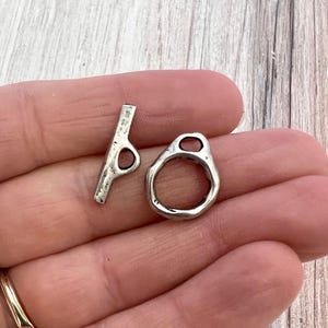 Könnte beinhalten: Ein silberfarbenes Knebelverschluss-Set, bestehend aus einem Stab mit einer kreisförmigen Öffnung und einem strukturierten Ring mit einer ovalen Öffnung. Der Verschluss ist für die Schmuckherstellung und das Basteln konzipiert.
