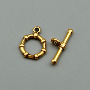 May include: A gold-toned toggle clasp set consisting of a circular ring with a small loop at the top and a matching T-bar component with a central loop. Both pieces feature a smooth, metallic finish with rounded, segmented detailing throughout.