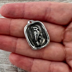 Puede incluir: Un colgante rectangular de tono plateado con un borde irregular estilo sello de cera. El centro presenta un relieve detallado de un pájaro posado en una rama. El colgante incluye un pequeño lazo circular en la parte superior.