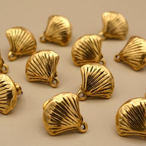 May include: A collection of gold-toned metal charms shaped like fan-like seashells. Each charm features a textured, ribbed surface and a small circular loop at the base for attachment. The pieces are arranged in a scattered pattern.