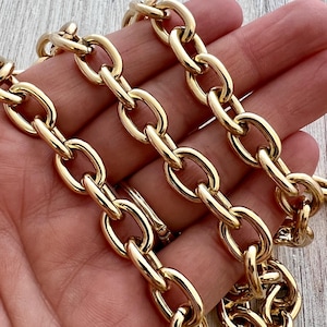 Puede incluir: Primer plano de un collar de cadena dorado con eslabones ovalados grandes. La cadena es gruesa y parece estar hecha de metal. Los eslabones están espaciados uniformemente y reflejan la luz, creando una apariencia brillante. El collar se sostiene en una mano.