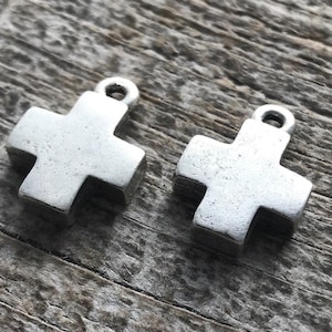 May include: Two silver-toned metal cross charms, each featuring a simple, thick, four-armed design with a small circular loop at the top for attachment. The charms have a matte, slightly weathered finish and a uniform, solid geometric shape.