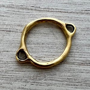 May include: A gold-colored, circular jewelry finding with two teardrop-shaped loops. The finding has a smooth, polished surface and is set against a light-colored, textured background. The design is simple and elegant.