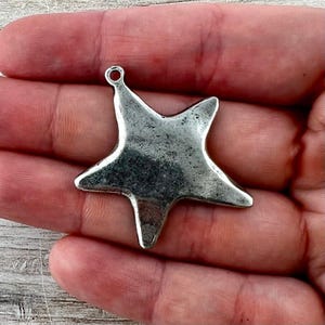 May include: A five-pointed star charm made of a metallic material with a weathered, dark silver finish. The star features a small circular loop at the top point for attachment, showcasing a textured, uneven surface across its entire front face.