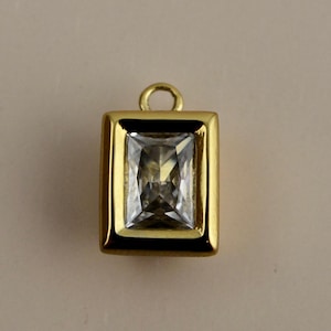 May include: A gold-colored pendant with a rectangular, clear gemstone. The gemstone is set within a gold frame, and the pendant has a small loop at the top for attaching to a chain or other jewelry.
