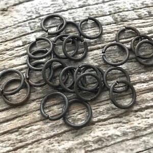 以下が含まれることがあります： 風化した木材の表面に散らばる、小さな濃いグレーの金属製丸カン。 リングは開いており、ジュエリー作りや工芸品に使用できます。 木目は素朴な雰囲気を添えています。