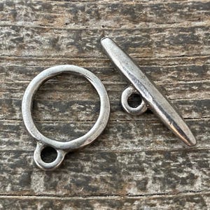 May include: A silver-toned toggle clasp, featuring a circular ring and a bar. The clasp is designed for jewelry making and is shown against a textured, wooden surface. The metal has a slightly aged appearance.