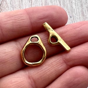 Könnte beinhalten: Zwei goldfarbene Knebelverschlüsse. Einer hat die Form eines Rings mit einer ovalen Öffnung, der andere ist ein Stab mit einer kreisförmigen Öffnung. Die Verschlüsse sind aus glänzendem Metall gefertigt, wahrscheinlich für die Schmuckherstellung oder Bastelarbeiten.