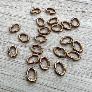 May include: A collection of small, gold-colored oval jump rings with a textured, rope-like pattern. The open rings are scattered on a light-colored wooden surface, ideal for jewelry making and crafting.