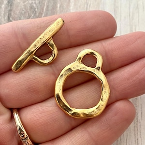 May include: Close-up of two gold-colored toggle clasps. One clasp is a bar with a loop, and the other is a ring with a loop. The clasps have a textured, hammered appearance. The clasps are likely for jewelry making or crafts.