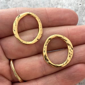 May include: Two gold-coloured, oval-shaped jewellery components with a textured, hammered appearance. The components are held in a hand, with a gold ring visible. The jewellery pieces have an organic, imperfect shape.