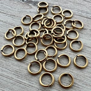 May include: A collection of numerous small, circular metal jump rings with an antique brass finish. Each ring features a single split opening, allowing them to be used as connectors for jewelry making, crafting, or various assembly projects.