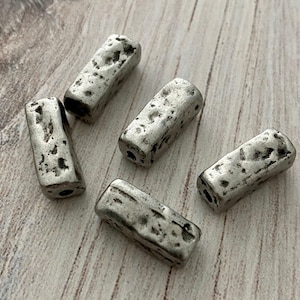 May include: Five rectangular beads with a metallic silver finish and a textured, pitted surface. Each bead features a hole running through the center, showcasing a rugged, uneven appearance across the entire exterior of the small metal components.