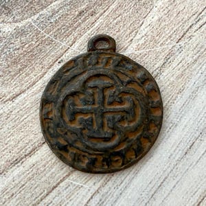 May include: A round, antique-style pendant with a cross design in the center. The pendant is a dark, weathered brown color, with a loop at the top for hanging. The design is embossed and detailed, giving it a vintage look.