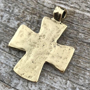 May include: A gold-toned metal cross pendant featuring a textured, uneven surface and a small loop at the top for attachment. The cross has wide, flared arms and a matte, weathered finish, presenting a solid, singular piece of jewelry hardware.