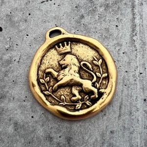 Puede incluir: Colgante dorado con diseño de león coronado, rodeado por una corona de laurel. El colgante tiene un lazo en la parte superior para una cadena.