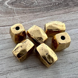 Könnte beinhalten: Sechs goldfarbene, facettierte Perlen mit gehämmerten Texturen. Jede Perle hat eine rechteckige Form mit einem Loch in der Mitte. Die Perlen sind auf einer hellen Holzoberfläche angeordnet.