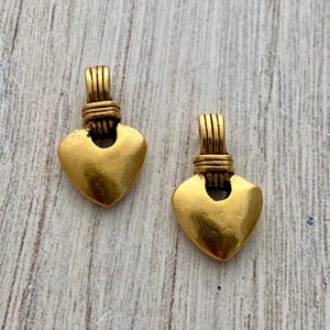 Può includere: Due ciondoli a forma di cuore in metallo color oro. Ogni ciondolo presenta una base a cuore liscia e solida fissata a un gancio verticale scanalato con dettagli orizzontali simili a fili metallici nel punto di giunzione.