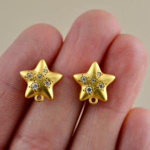 May include: Two gold-toned star-shaped charms with a matte finish, each featuring five small, clear, round-cut embedded stones arranged in a cluster. Each star charm includes a small circular loop at the bottom for attachment to jewelry.