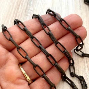 May include: Close-up of a dark brown metal chain with oval links. The chain is arranged in parallel lines, with a slight curve. The metal has a rustic, aged appearance. A gold ring is visible in the lower left corner.