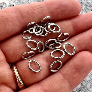 May include: A collection of small, silver-colored oval jump rings with a textured, twisted rope design. The rings are held in a hand, showcasing their size and detail. The rings are for jewelry making.