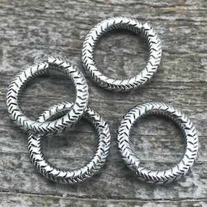 May include: Four silver-colored, textured metal rings with a repeating chevron pattern. The rings are arranged on a weathered, gray wooden surface, creating a rustic aesthetic. These rings could be used for jewelry making or other crafts.