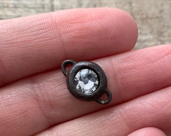 Könnte beinhalten: Ein kleines, oval geformtes Metallverbindungsstück mit einem dunklen, verwitterten Bronzefinish. In der Mitte befindet sich ein einzelner, klarer, facettierter runder Kristallstein. Das Stück hat zwei kleine kreisförmige Ösen an den Enden.