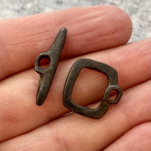 May include: A dark metallic toggle clasp set consisting of a T-shaped bar with a central hole and a square-shaped loop with an integrated eyelet. The components feature a weathered, matte finish with visible surface texture and dark patina.