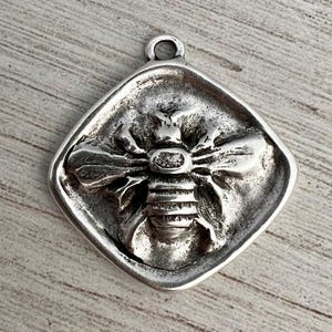 以下が含まれることがあります： 正方形のフレーム内に詳細な蜂のデザインが施されたシルバーカラーのペンダント。 蜂は、複雑な翼と体のセグメントで描かれています。 ペンダントの上部には吊り下げ用のループがあり、おそらく金属製です。