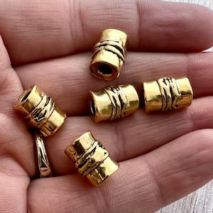 May include: Five cylindrical gold-toned beads with a textured, organic surface featuring horizontal ridges and a central hole. Each bead has a polished metallic finish and a slightly irregular, hand-crafted appearance, arranged in a cluster.