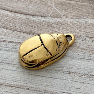 May include: A gold-coloured scarab beetle pendant with a loop for hanging. The beetle has detailed markings and a textured surface, suggesting an antique or vintage style. The pendant is likely made of metal.