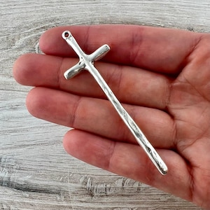 Puede incluir: Un colgante de cruz plateada con un acabado texturizado. La cruz tiene un diseño tradicional con una barra vertical larga y una barra horizontal más corta. Cuenta con un pequeño lazo en la parte superior para colgar.