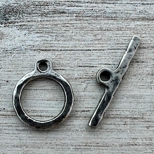 Puede incluir: Un juego de cierre de palanca plateado, que presenta un anillo con un lazo y una barra con un lazo. El cierre tiene un aspecto rústico y texturizado. Ideal para la fabricación de joyas, este cierre se muestra sobre un fondo de vetas de madera clara.