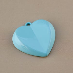May include: A pale blue heart-shaped decoration with a loop for hanging. The heart has a shiny surface and a faceted design. The background is a neutral beige colour.