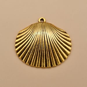 May include: A gold-toned metal pendant shaped like a scallop shell. The surface features a series of raised, radiating ridges that extend from the top center to the scalloped bottom edge. A small circular loop is attached at the top for hanging.