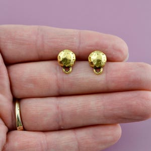 May include: Two small, gold-colored stud earrings. Each earring has a textured, spherical design with a small loop at the bottom. The earrings are displayed against a purple background.