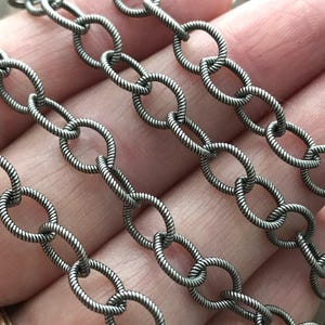 May include: Close-up of a silver chain with oval links. The chain has a textured, twisted rope design. The links are connected in a repeating pattern. This chain is suitable for jewellery making.
