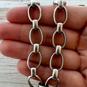 May include: Close-up of a silver-toned chain bracelet. The bracelet features large oval links connected by rectangular bars. The metal has a slightly aged appearance, suggesting a vintage or antique style. The design is simple yet bold, suitable for various styles.