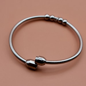 May include: A silver-toned metal bracelet featuring a flexible snake chain design. The bracelet includes three small spherical beads positioned along the top section and two cylindrical end caps that meet at the opening of the circular band.