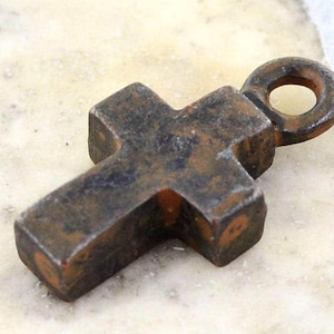 May include: A small, dark, weathered metal cross pendant with a thick, blocky shape and a textured, rusted surface. The cross features a circular loop at the top for attachment, displaying a worn, aged appearance with mottled brown and black tones.