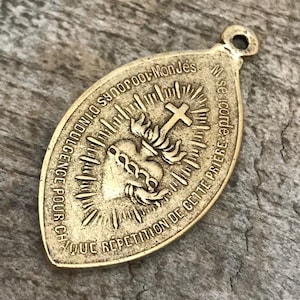 Puede incluir: Colgante ovalado dorado con un diseño religioso. El colgante presenta un corazón con llamas, una cruz y texto en francés. Tiene un pequeño lazo en la parte superior para sujetarlo a una cadena o cordón.