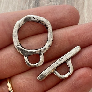 May include: A silver-toned metal toggle clasp set consisting of a circular ring component with a small loop attachment and a straight bar component with a central loop attachment, both featuring a textured, weathered, and rustic metallic finish.