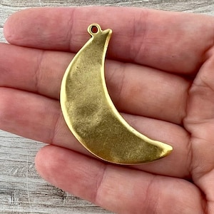 May include: A brass-colored crescent moon pendant with a textured surface. The pendant has a small loop at the top for attaching to a chain or cord. The moon shape is curved and detailed, suggesting a celestial or bohemian theme.