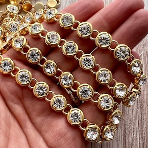 May include: Close-up of gold-toned jewelry featuring clear, round gemstones set in circular bezels. The gemstones are linked together with small gold connectors. The jewelry is displayed on a light-colored surface.