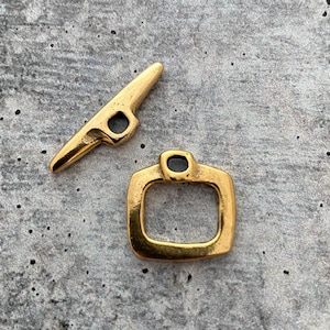 May include: A gold-toned metal toggle clasp set consisting of a T-shaped bar with a central loop and a matching square-shaped ring with a small attachment point. The pieces feature a smooth, polished finish with subtle variations in the metal surface.