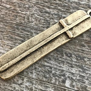 May include: A rectangular, antique brass-colored pendant with a cross design. The pendant has a loop at the top for attaching to a chain or cord. The cross is raised and centered on the pendant.