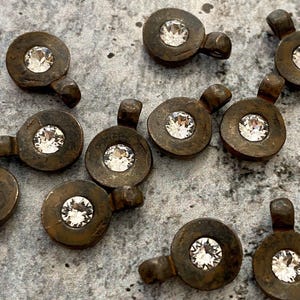 May include: Close-up of several antique bronze-colored metal charms. Each charm features a circular design with a clear, faceted crystal in the center and a small loop. The charms are scattered on a textured, gray surface.